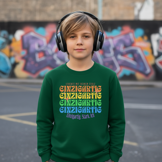Junge mit Niose-cancelling-Kopfhörern trägt grünes Sweatshirt mit regenbogenfarbenem Schriftzug „Einzigartig. Stark. Ich.“ – liebevolles Statement-Design für Kinder, das Vielfalt und Selbstliebe zeigt.